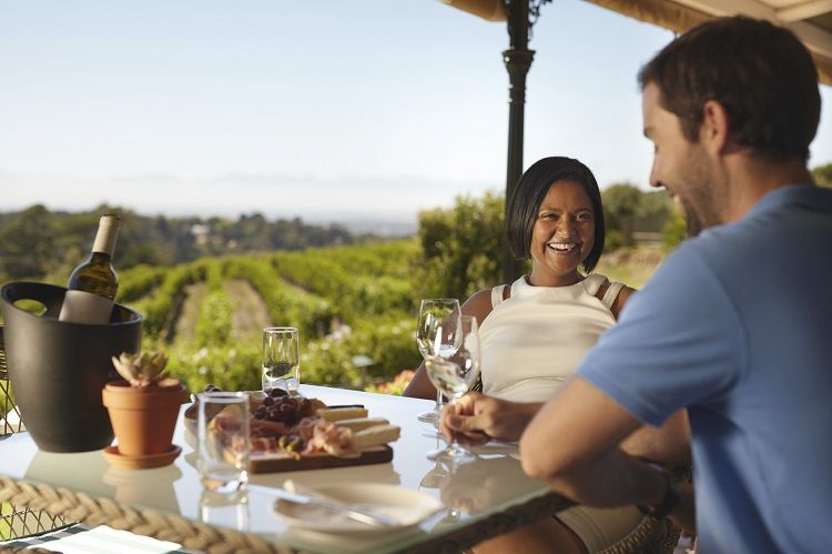 Happy young couple at winery restaurant with vineyard in background. Smiling young woman with a man sitting at table drinking wine.
