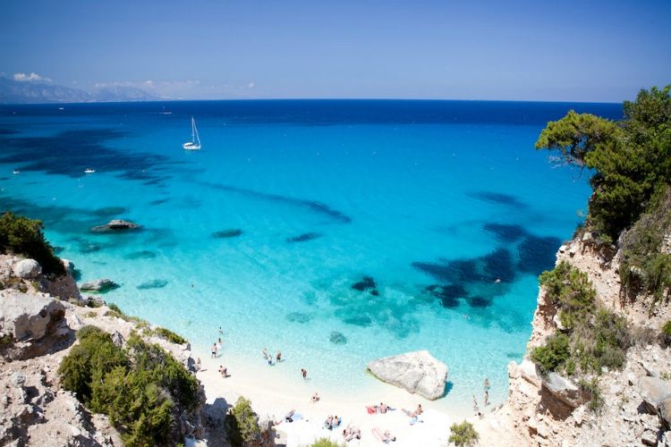 beaches in sardinia | drone view of white sand beach and blue water with sailboat under clear skies | Cala Goloritzè