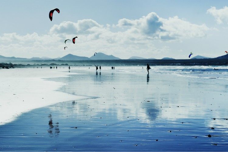 a panoramic view of the Famara Beach in Lanzarote, Canary Islands, Spain, with some unrecognizable swimmers and kitesurfers in the background, in backlit (a panoramic view of the Famara Beach in Lanzarote, Canary Islands, Spain, with some unrecognizab